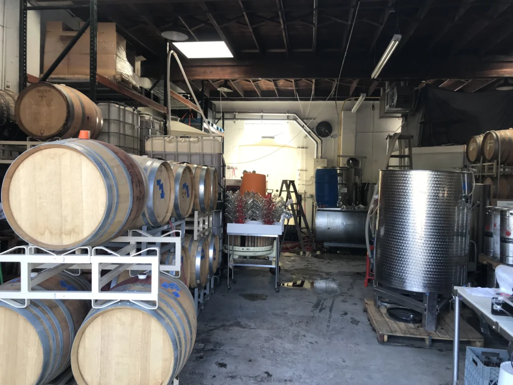 The working cellar: oak barrels racked along one wall, totes and a stainless tank opposite, hoses and empty bottles waiting between them.